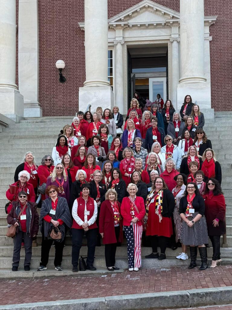 Large group of WCRW members wearing red scarves posed on the steps of a brick building with tall white columns during Red Scarf Day 2025