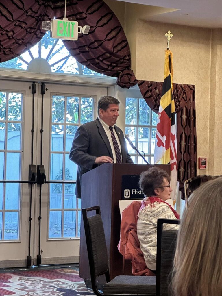 Maryland State Delegate Wayne A. Hartman (District 38C) speaks at a podium with the Maryland state flag behind him during the Red Scarf Day 2025 event