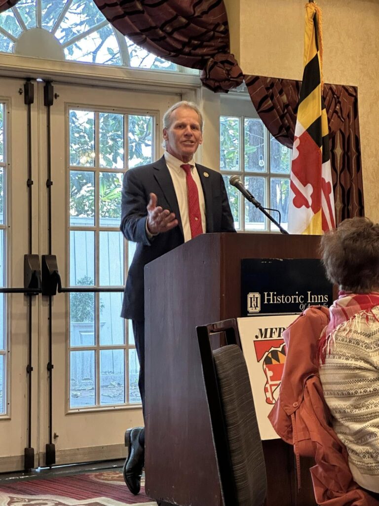 Maryland State Delegate Barry S. Beauchamp (District 38B) speaks at a podium with the Maryland state flag behind him during the Red Scarf Day 2025 event
